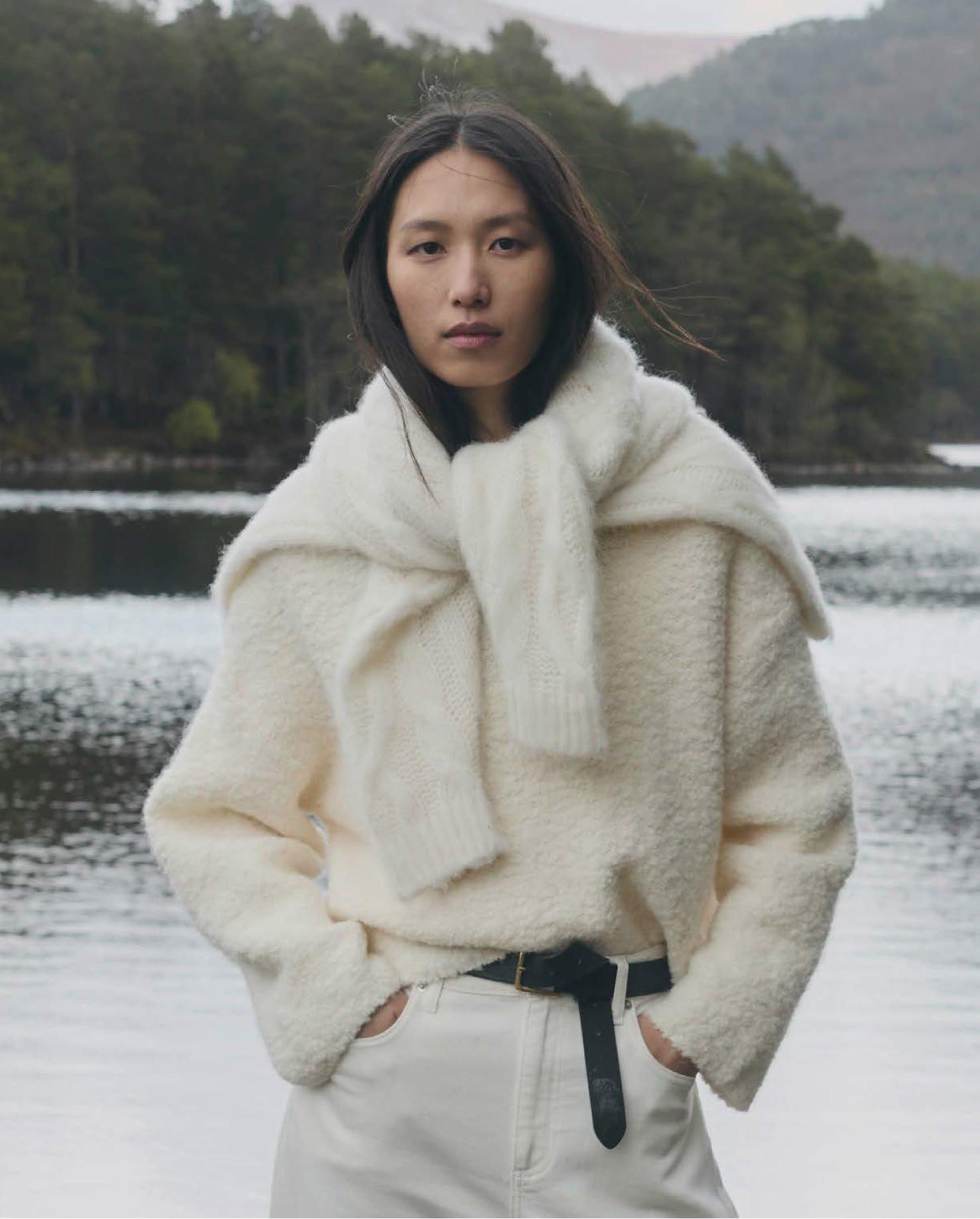 a woman in a white sweater and pants standing by a lake