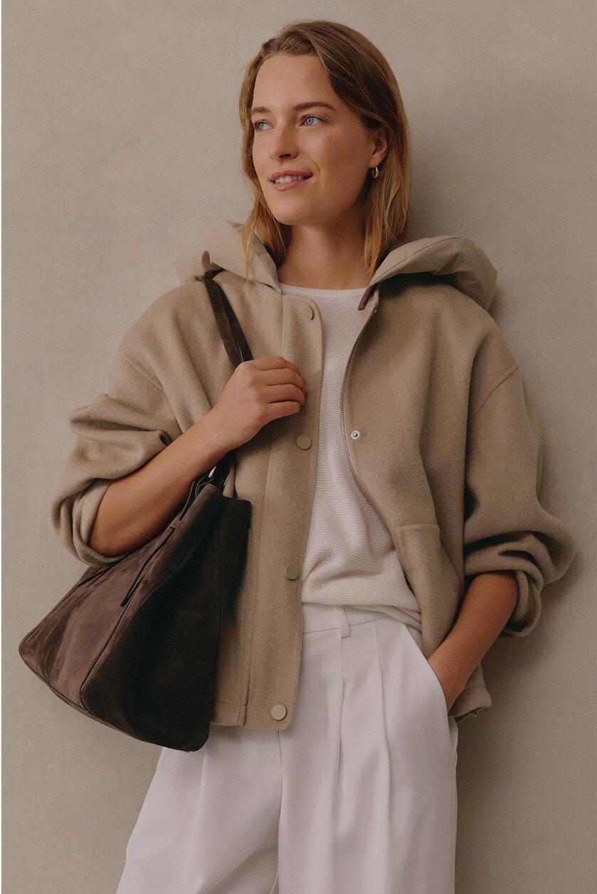 a woman in white pants and a beige jacket holding a brown bag