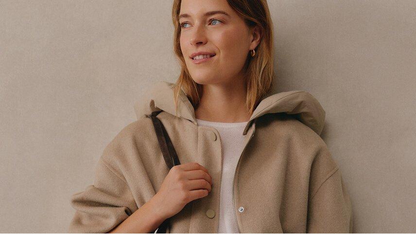 a woman in white pants and a beige jacket holding a brown bag