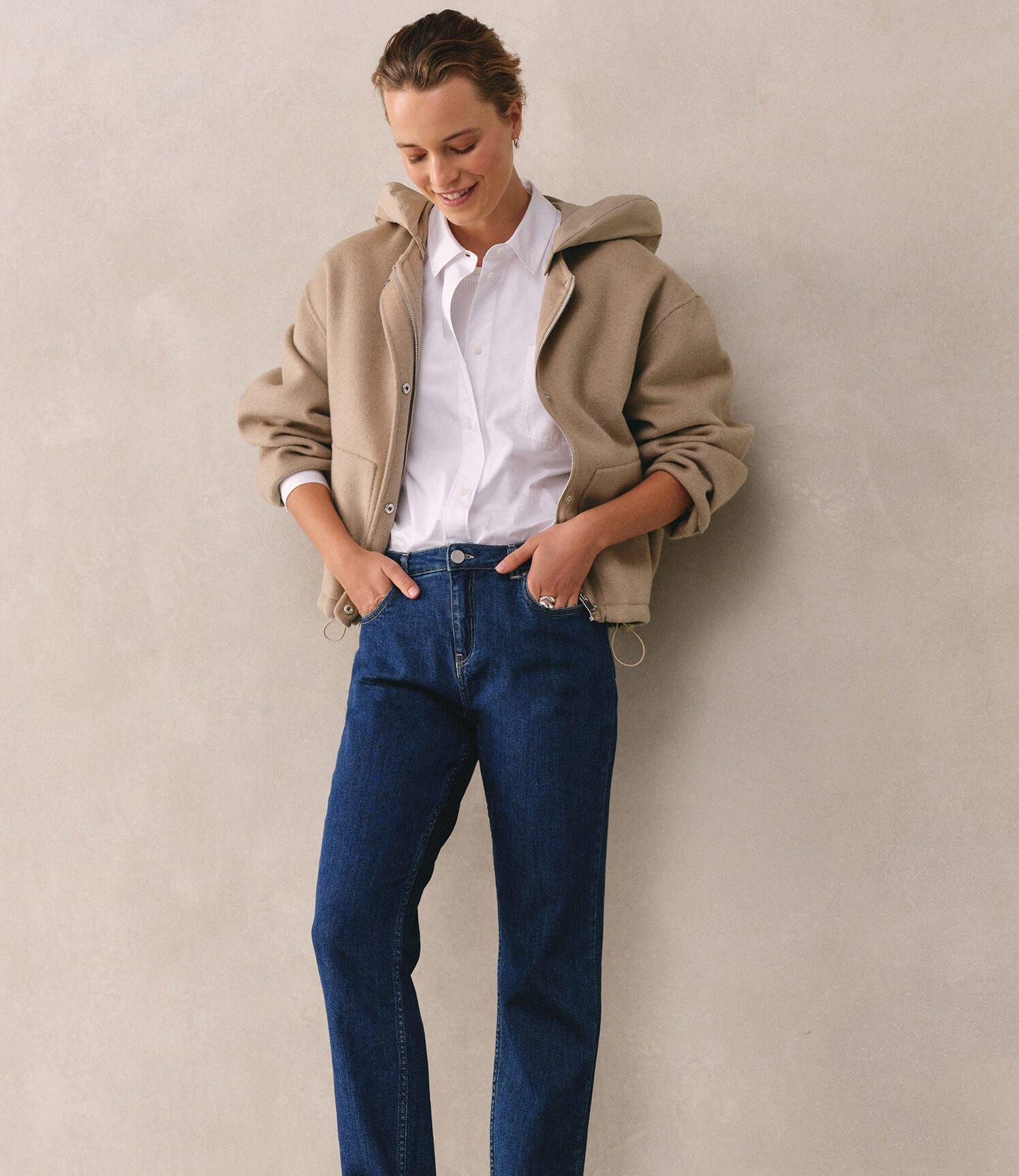 a woman in jeans and a jacket leaning against a wall