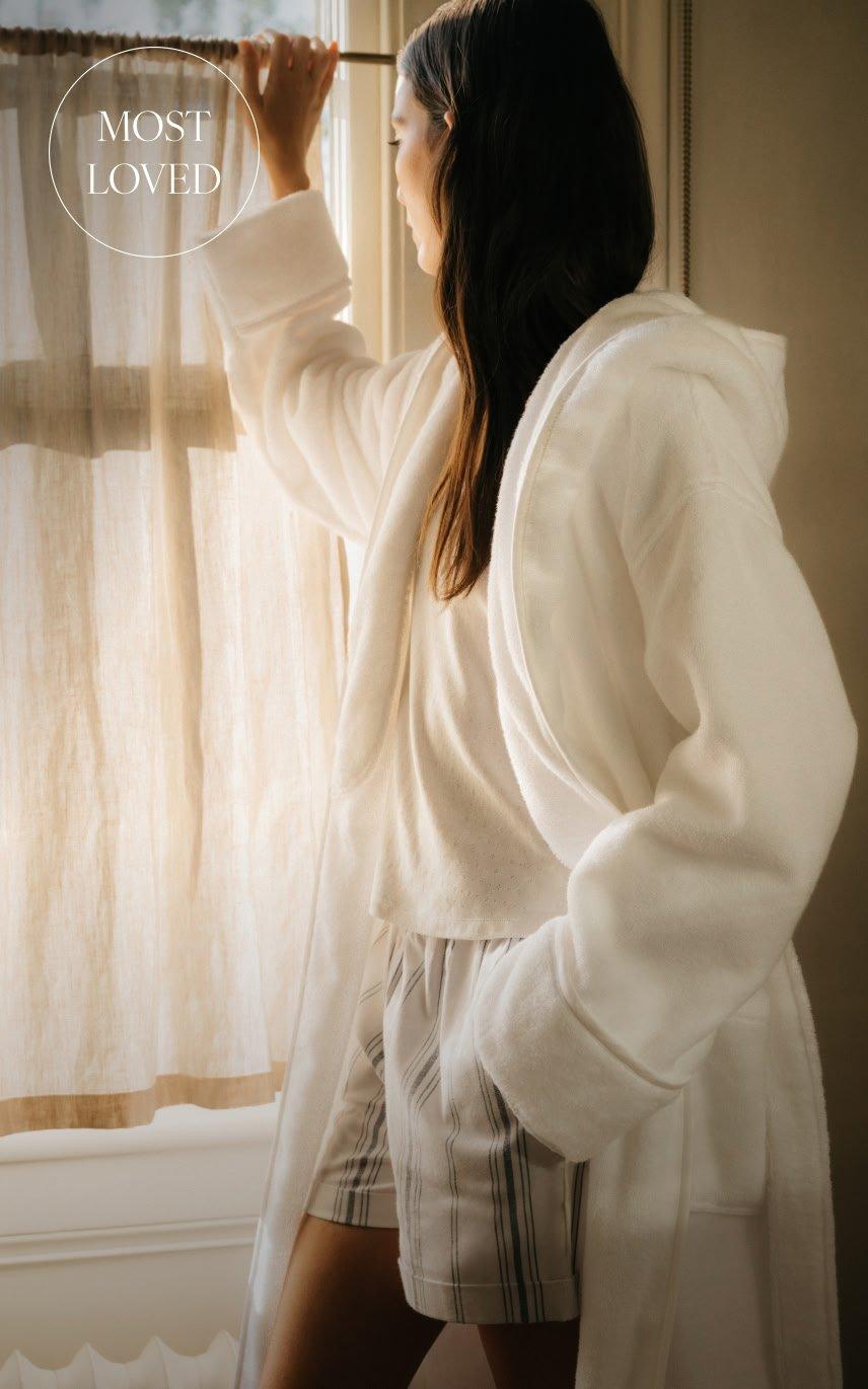a woman in a white robe looking out the window at a curtain