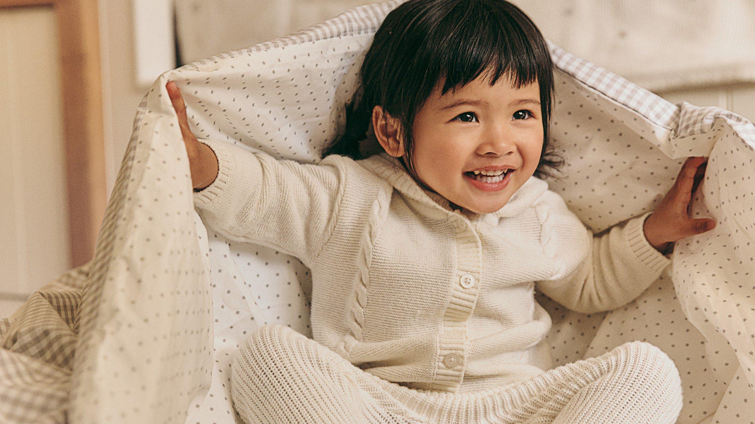 a little girl in white sitting on a bed with a blanket