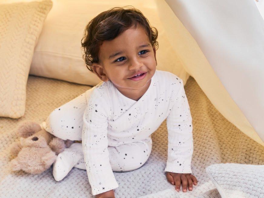 a baby in white pajamas sitting on a bed with a teddy bear