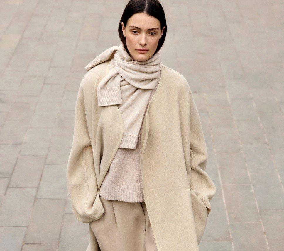 a woman in a beige coat and sweater walking down a street