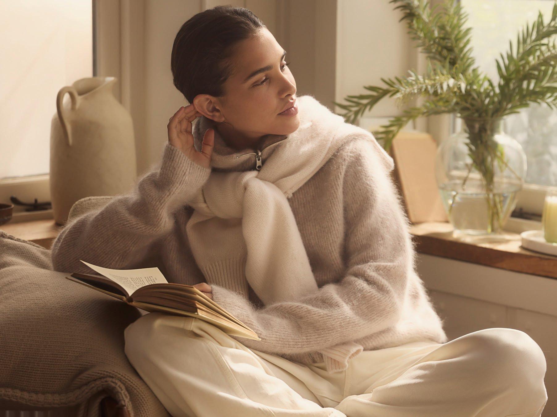 a woman in a sweater and scarf reading a book on a chair