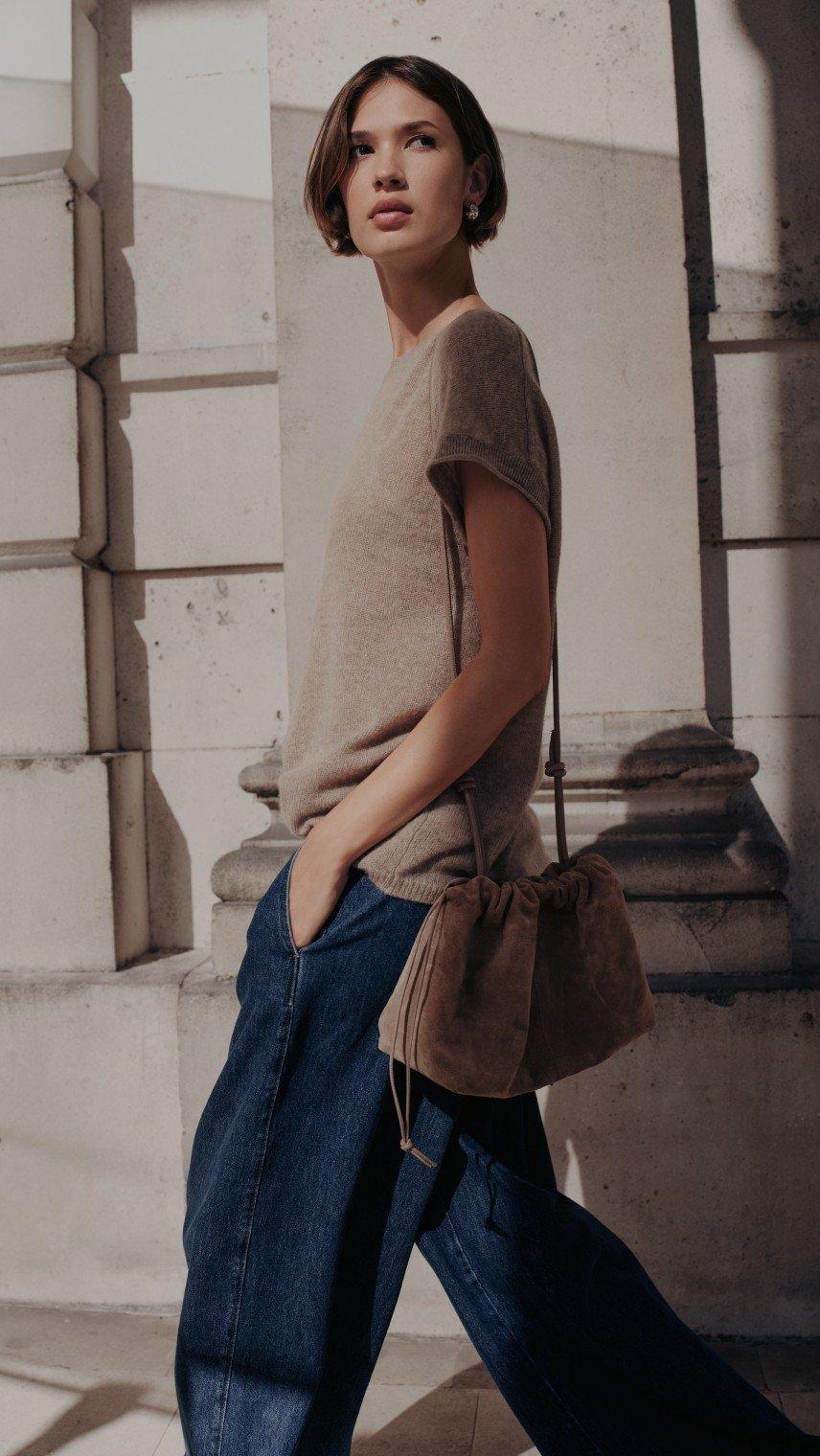 a woman in a beige sweater and brown purse standing next to a wall