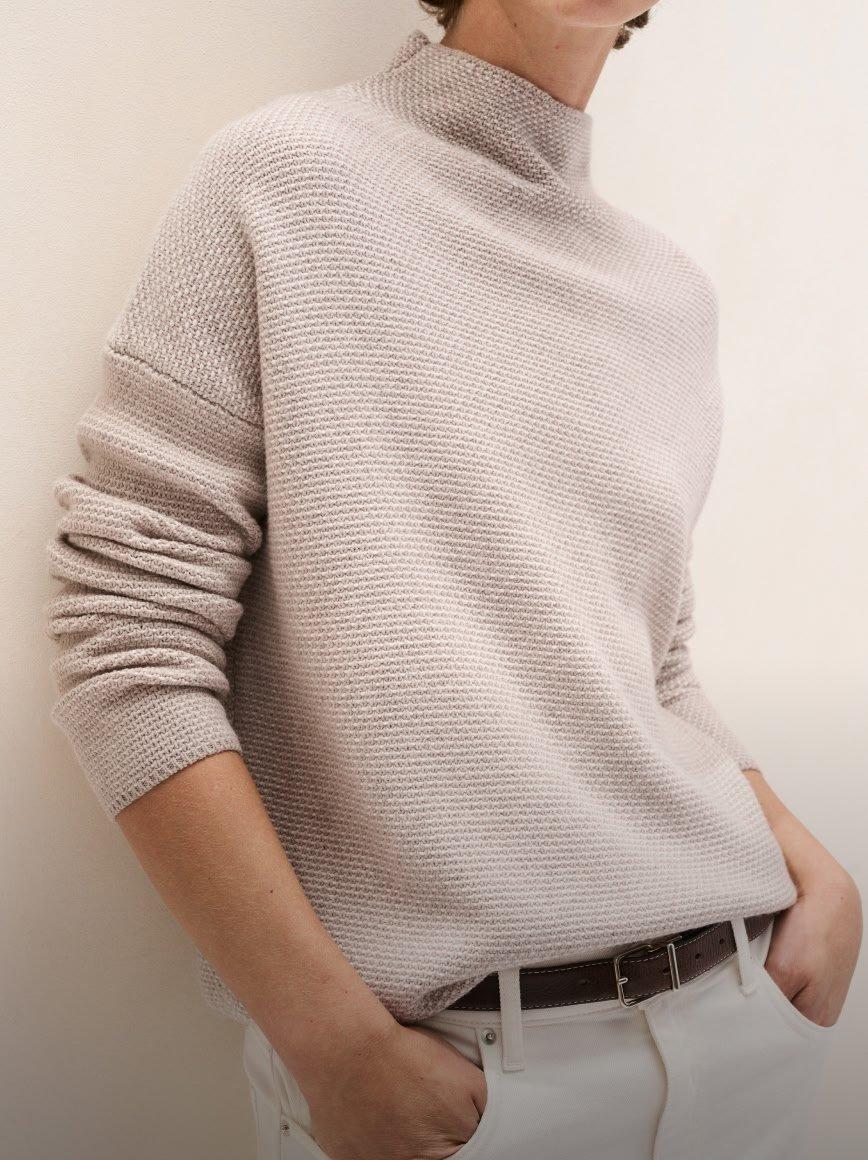 a man in a sweater and white pants leaning against a wall
