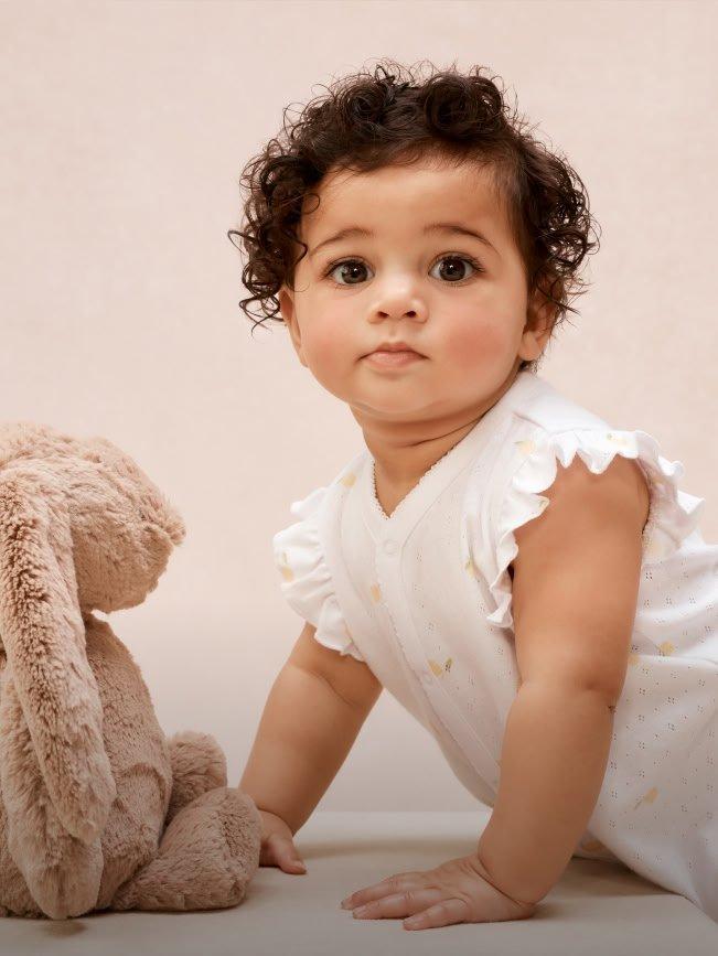 a baby girl with white shirt and white pants standing next to a stuffed animal