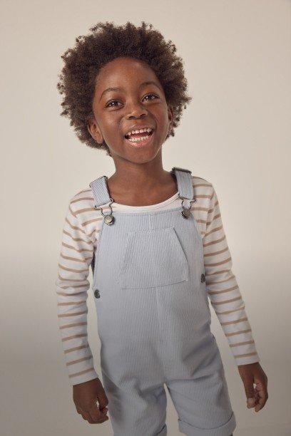a young boy in overalls smiling while holding a skateboard