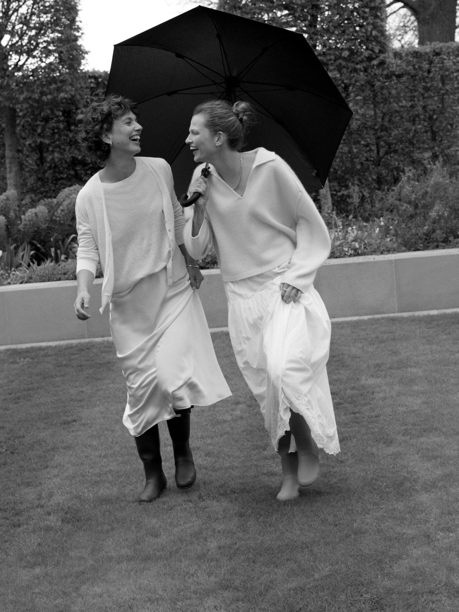 two women in white dresses holding an umbrella walking in a field