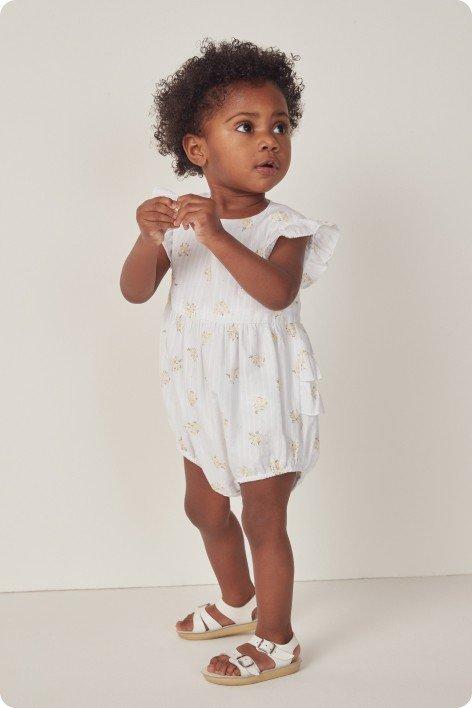 a toddler in a white romper with sandals standing on a white floor